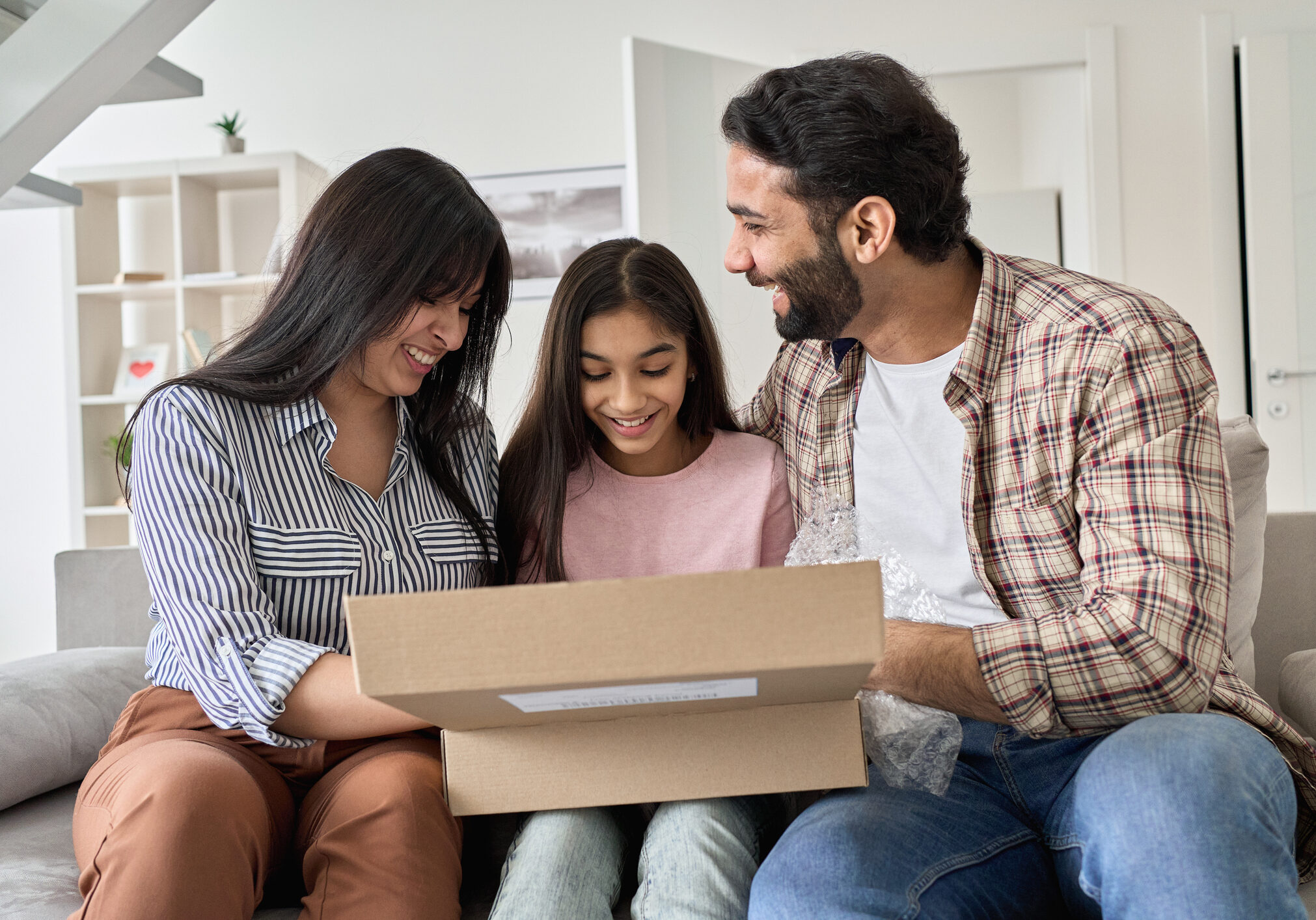 Happy indian family with child daughter unpacking parcel at home. Smiling parents and teen kid daughter opening postal box looking at gift in online shopping delivery package sitting on sofa together.